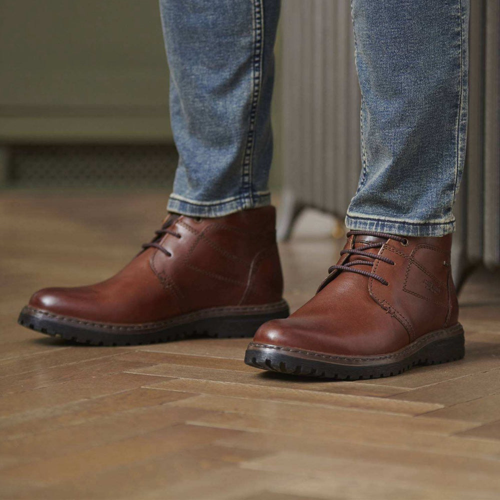 A pair of men's brown leather lace-up ankle boots with rugged black soles, shown on a person wearing blue jeans standing on a wooden floor.