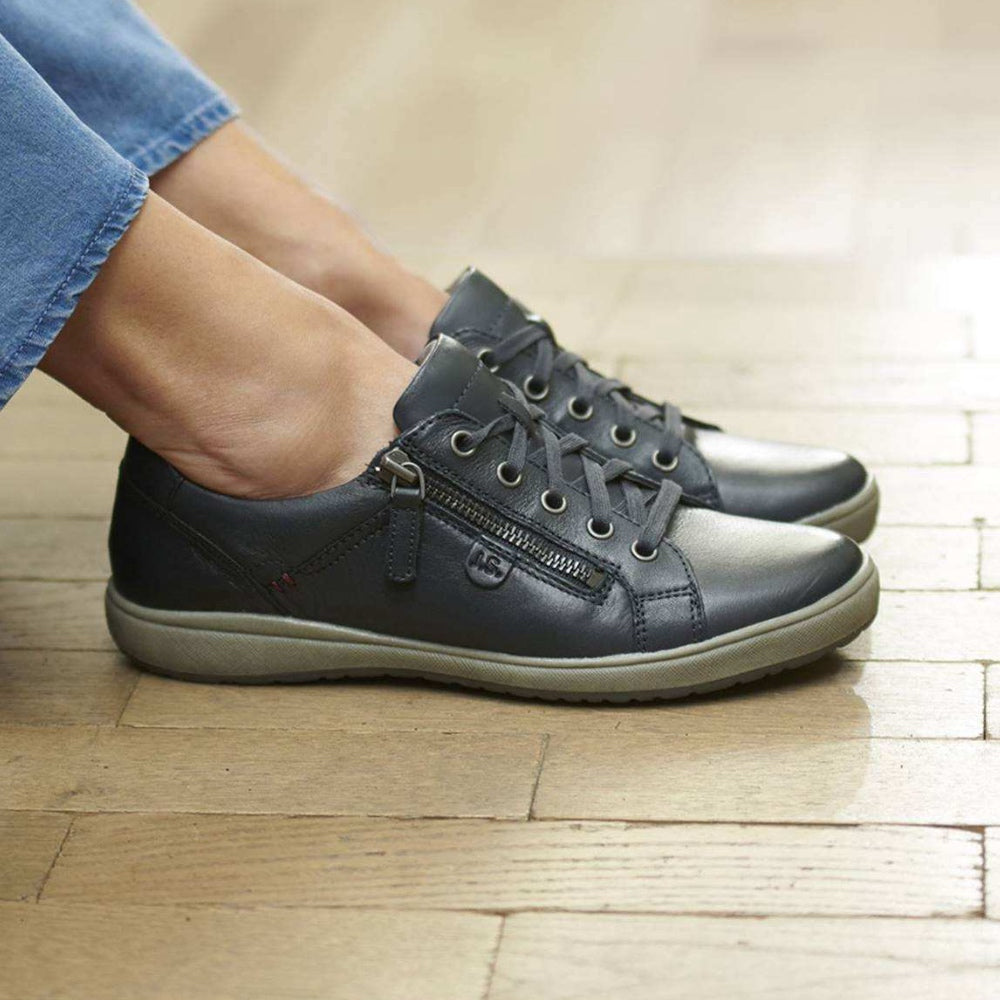 A pair of dark gray leather low-top sneakers with side zippers and lace-up fronts, shown being worn with blue jeans on a light wood floor.