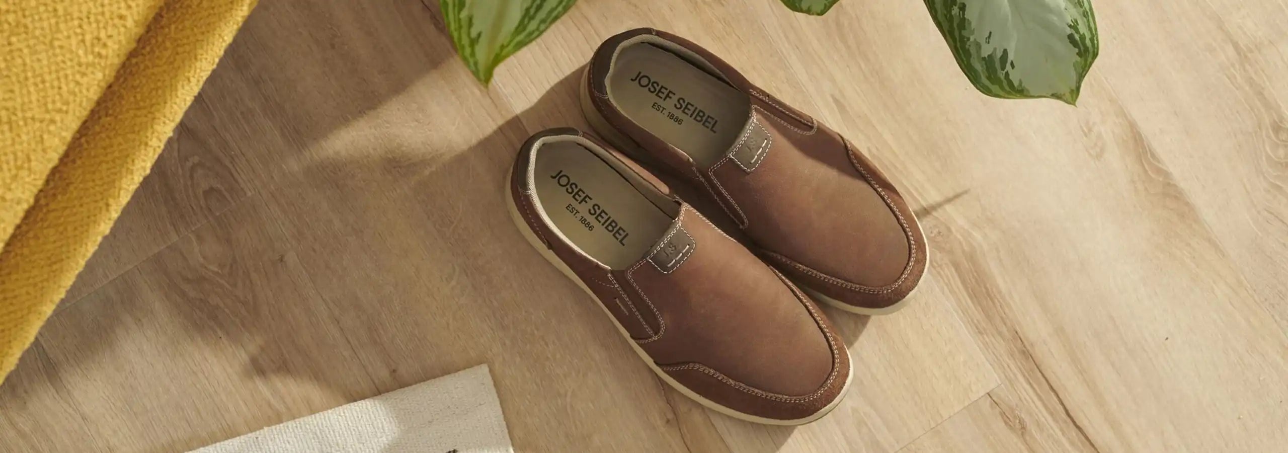 Brown slippers on a wooden floor with a yellow rug and green plants in the background