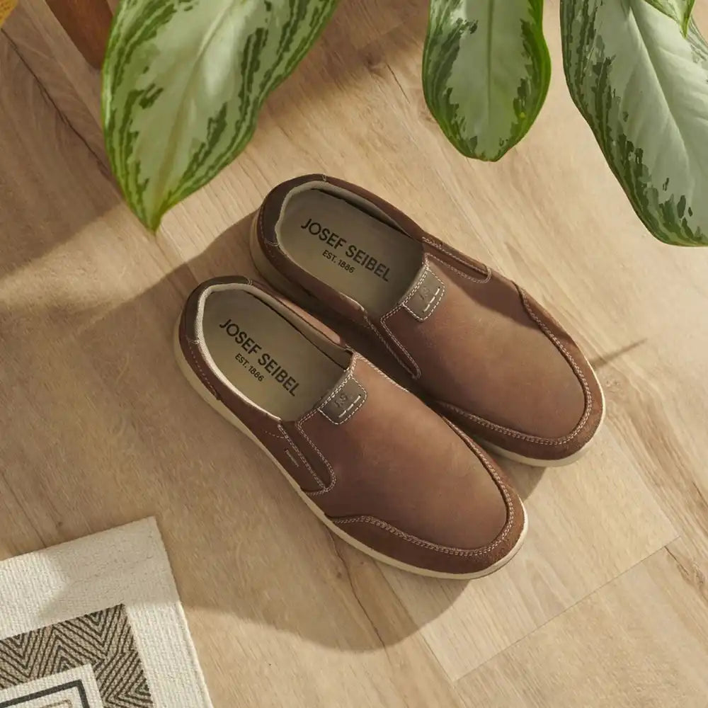 Brown loafers with 'Josef Seibel' branding on a wooden floor with plants in the background.