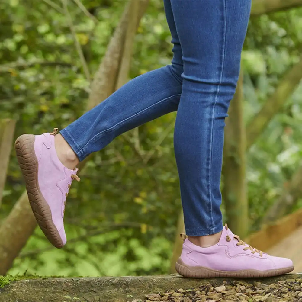 Person wearing pink shoes with brown soles and blue jeans, standing on a wooden platform with a green, natural background.